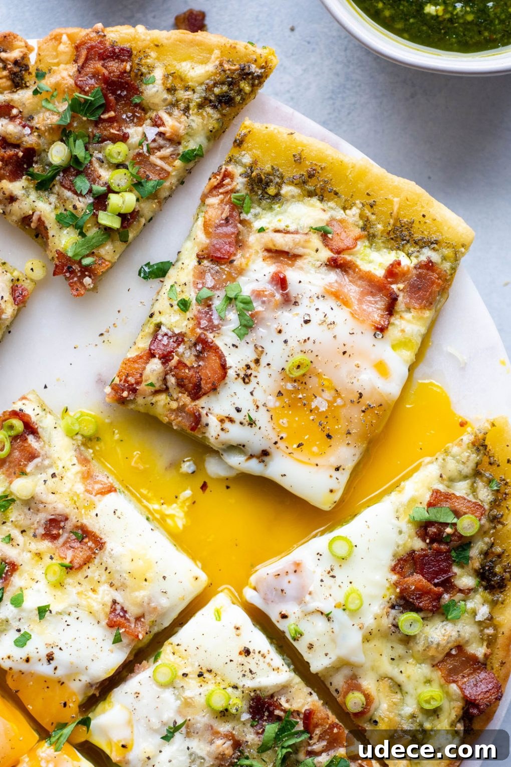 5-Ingredient Breakfast Pizza with Pesto and Crispy Bacon 3 A close up overhead shot of a single square shaped piece of breakfast pizza pulled away from the rest of the pizza surrounded by runny egg yolk and topped with fresh herbs and green onion.