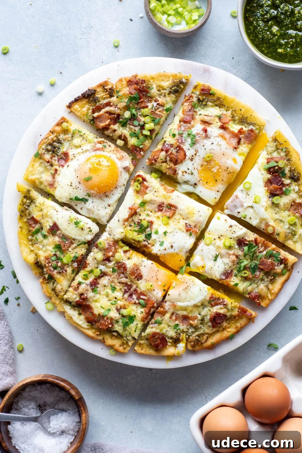 5-Ingredient Breakfast Pizza with Pesto and Crispy Bacon 4 Overhead shot of a breakfast pizza cut into 9 squares topped with melted cheese, crispy bacon, sunny side up eggs, and fresh herbs.