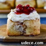 A front view of a cinnamon roll with cherry toppings on a wooden board with three cinnamon rolls in the background.