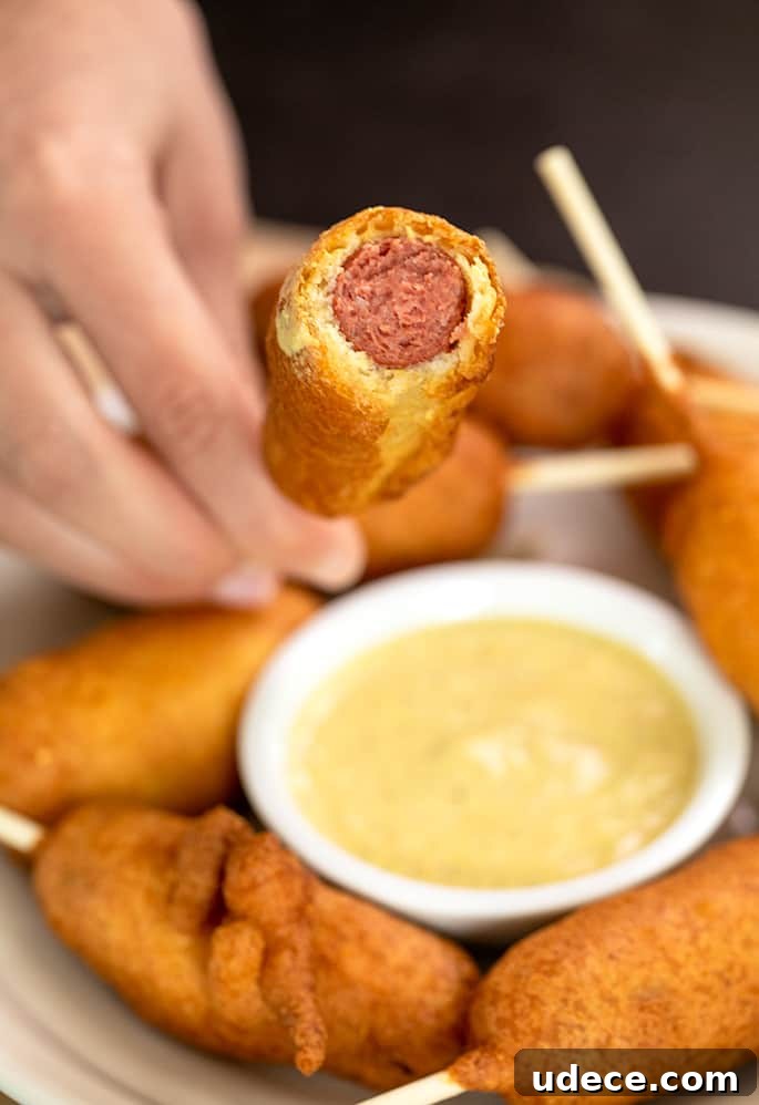 hand holding corn dog stick with end bitten off to reveal hot dog inside, over small bowl of mustard
