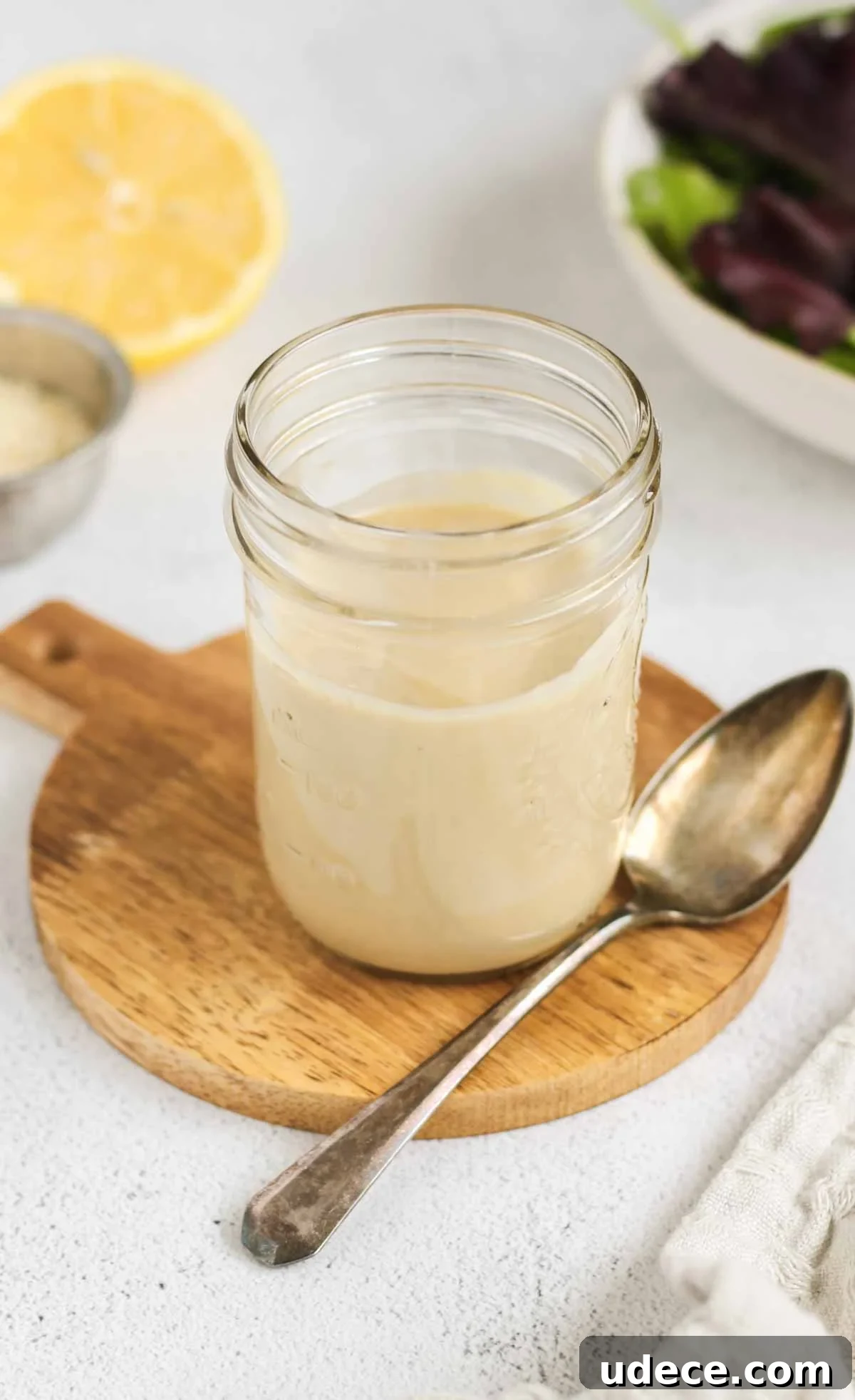 Maple Tahini Dressing Recipe for Salads and Bowls 2 Maple tahini dressing in a mason jar with a spoon on the side of the jar and a lemon and bowl of mixed greens in the background.