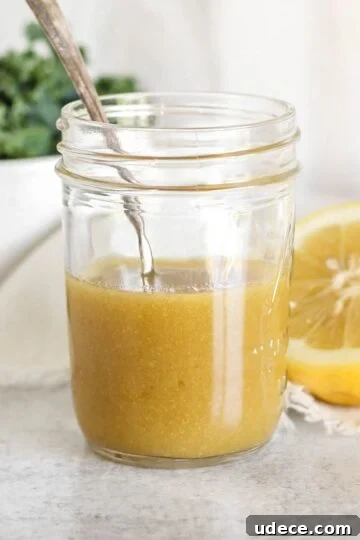 Maple Dijon dressing and a spoon in mason jar with a bowl of greens and a sliced lemon on the background.