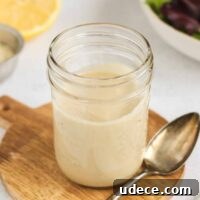 Maple tahini dressing in a mason jar with a spoon on the side of the jar and a lemon and bowl of mixed greens in the background.