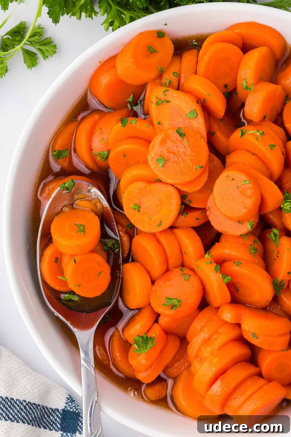 Overhead view of a serving bowl with brown sugar and honey glazed canned carrots and a spoon resting inside, garnished with chopped parsley.