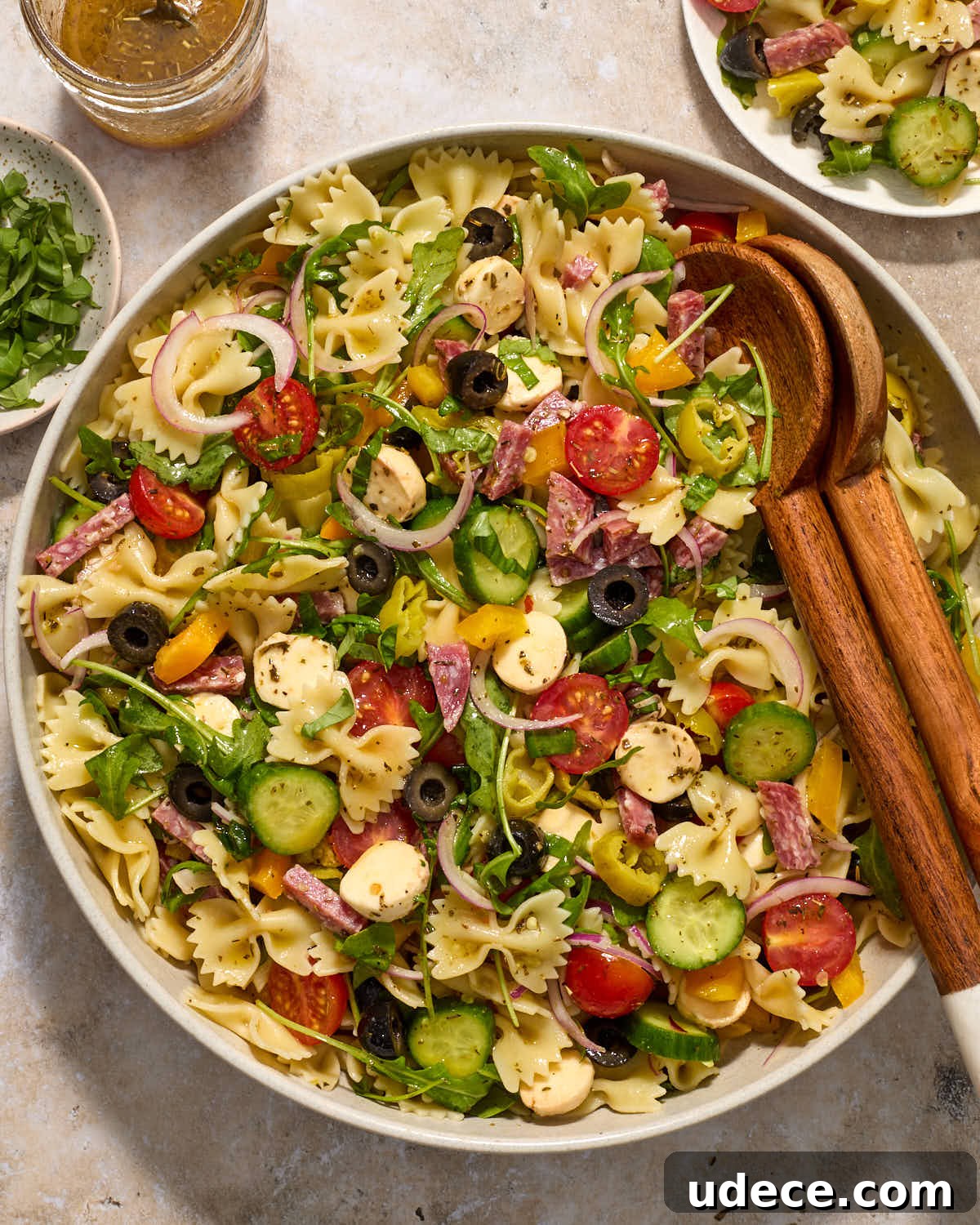 Pasta salad in a bowl with a plate of salad, extra vinaigrette and basil on the side.