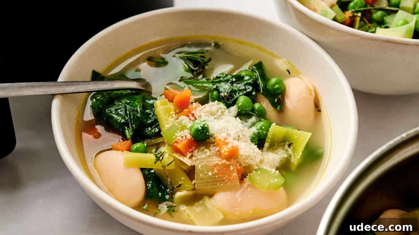 A bowl of vegetable soup with spinach, peas, carrots, celery, and large white beans, topped with grated cheese.