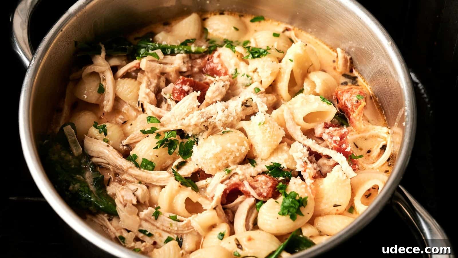 A pot of pasta with shredded chicken, spinach, tomatoes, and grated cheese, garnished with chopped parsley.