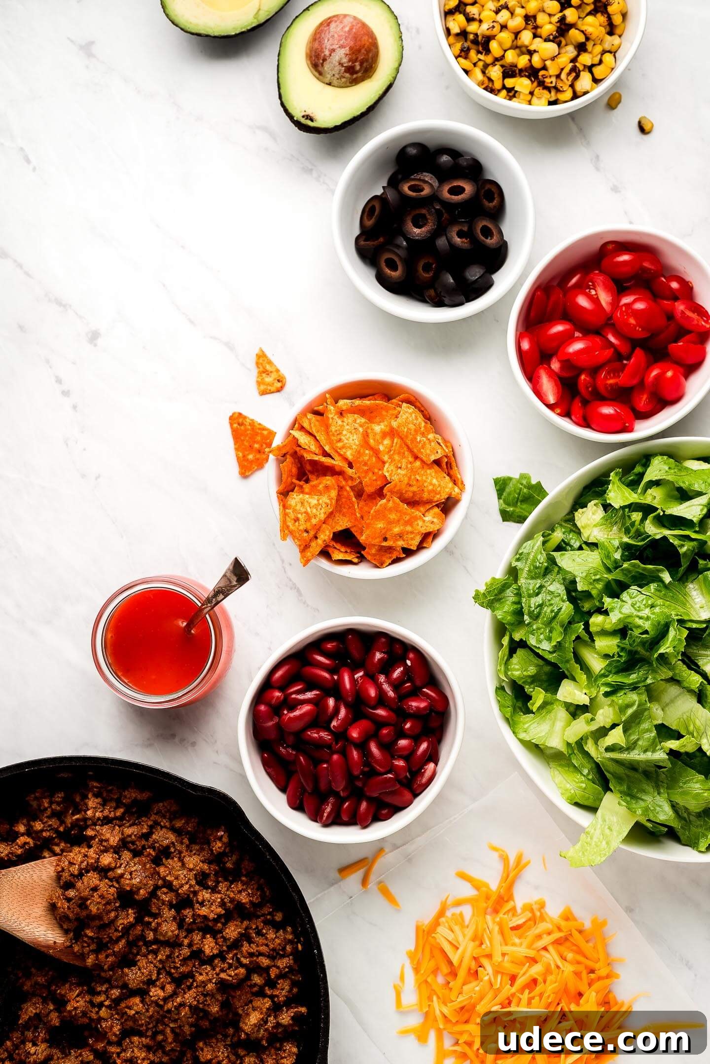 Ingredients on a marble surface- roasted corn, avocado, black olives, tomatoes, Doritos, lettuce, kidney beans, shredded cheese, taco meat, and dressing.