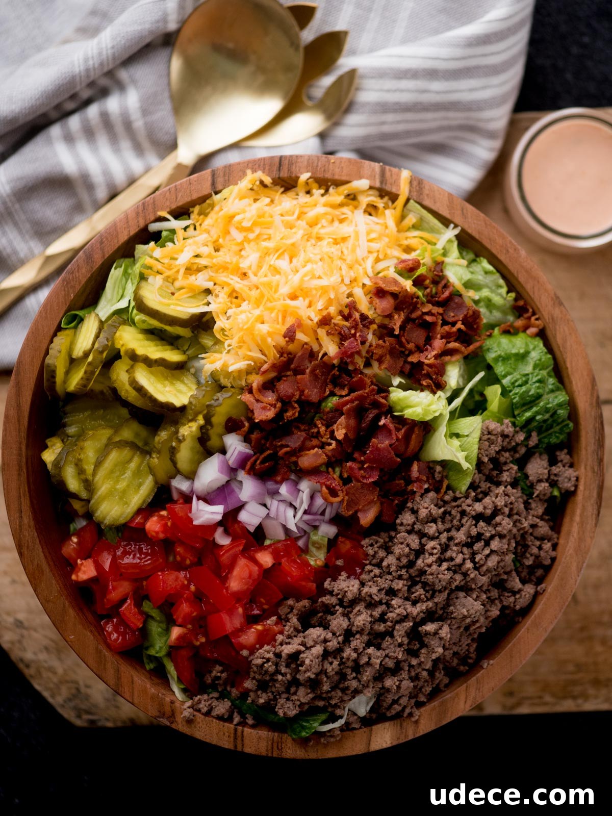 Copycat Big Mac Salad Recipe for Classic Burger Flavor 2 big mac salad in a wooden bowl sitting on a dough board. There are serving utensils, a jar of dressing, and a kitchen towel surrounding it.