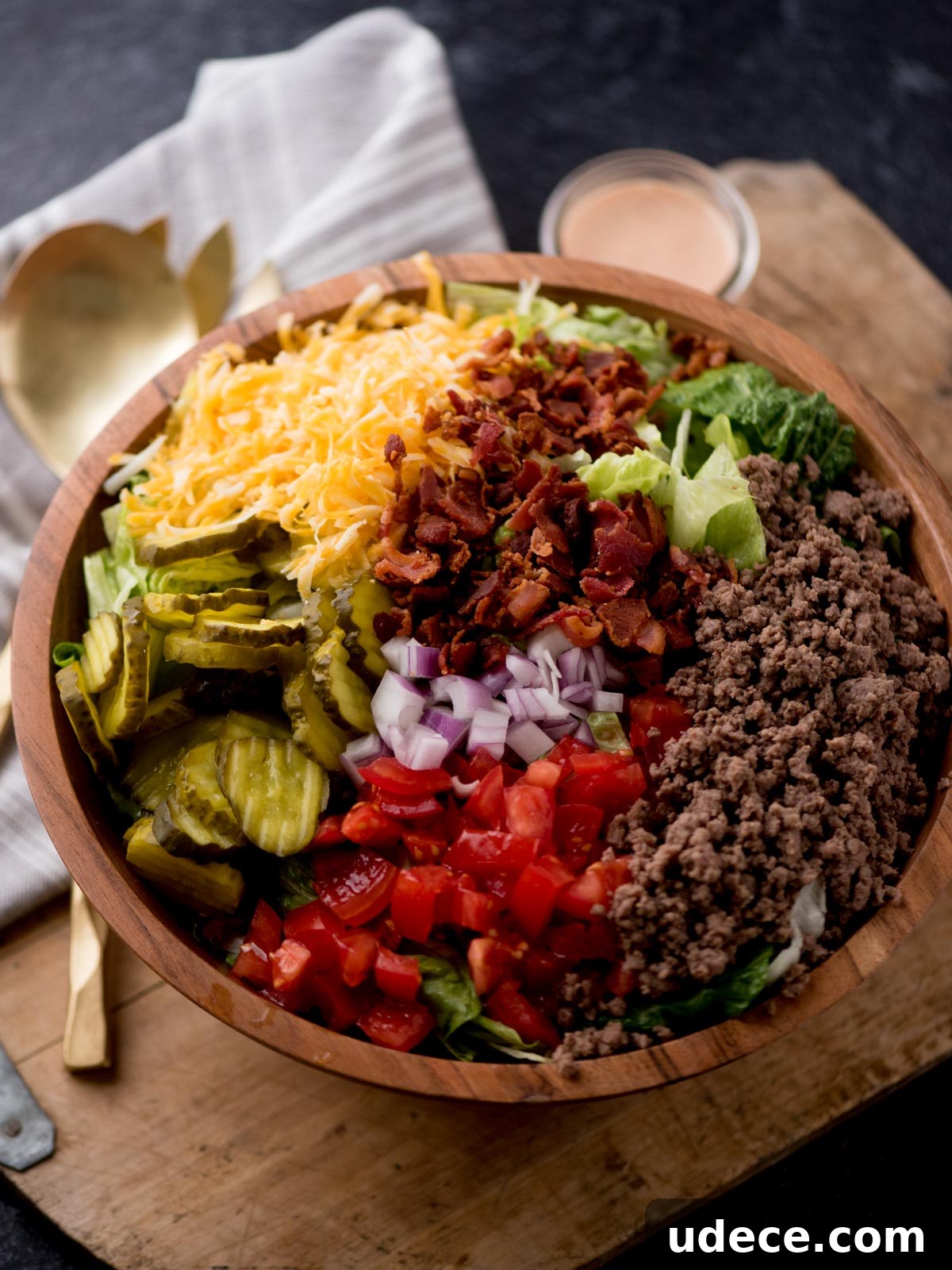 Copycat Big Mac Salad Recipe for Classic Burger Flavor 6 big mac salad in a wooden bowl sitting on a dough board. There are serving utensils, a jar of dressing, and a kitchen towel surrounding it.