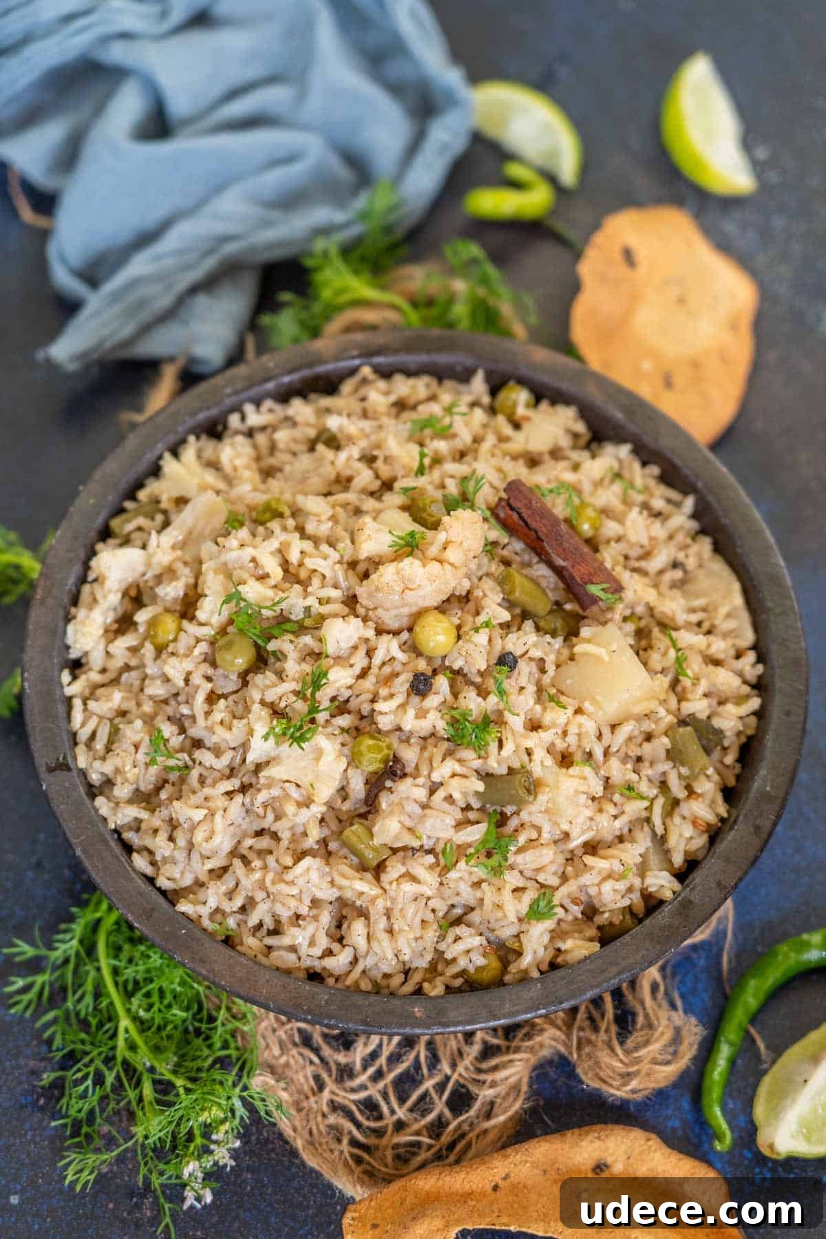 Flavorful Brown Rice Pulao: Aromatic One-Pot Indian Rice Recipe 2 A bowl of Brown Rice Pulao garnished with fresh herbs, featuring visible peas, cauliflower, and a cinnamon stick, surrounded by papad, lime wedges, green chili, and a blue cloth on a dark surface.