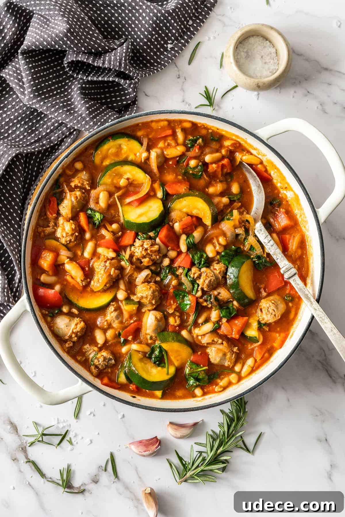 Mediterranean Sausage and Vegetable Casserole Recipe 2 Large saucepan of Sausage Casserole with a spoon in the pan, sitting on a marble bench top with some herbs around the edge.