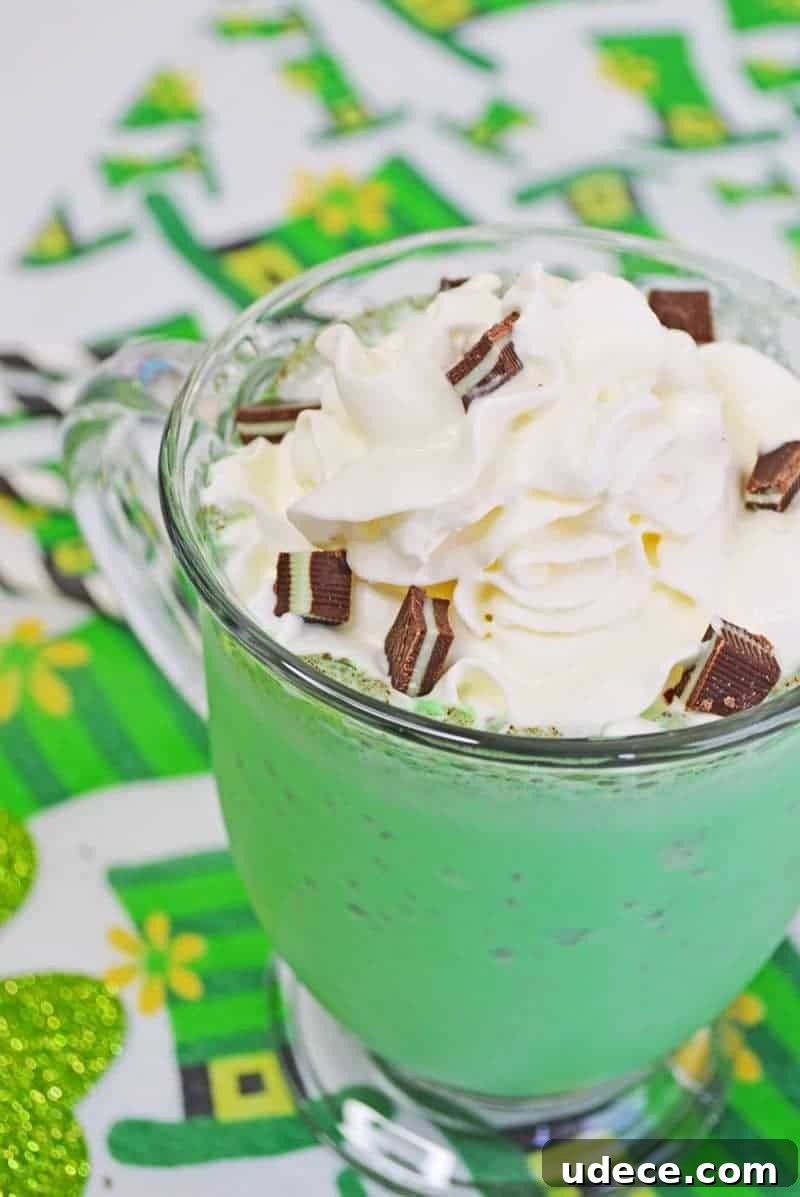 Shamrock Shake Recipe: Refreshing Minty Milkshake for St. Patrick’s Day 3 Reads: Shamrock Shake. Green mint chocolate chip milkshake in glass mug with whipped cream, chopped Andes Mints, and a white and black striped paper straw. The mug sits on a leprechaun hat tablecloth.