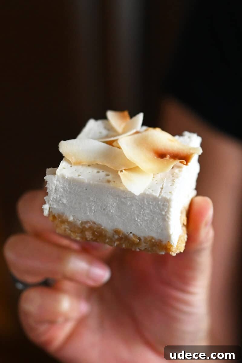 Vegan Coconut Cream Bars — Low-Carb, Gluten-Free Dessert 2 A closeup of a hand holding square of vegan, paleo, and gluten-free coconut cream bar topped with toasted coconut flakes.