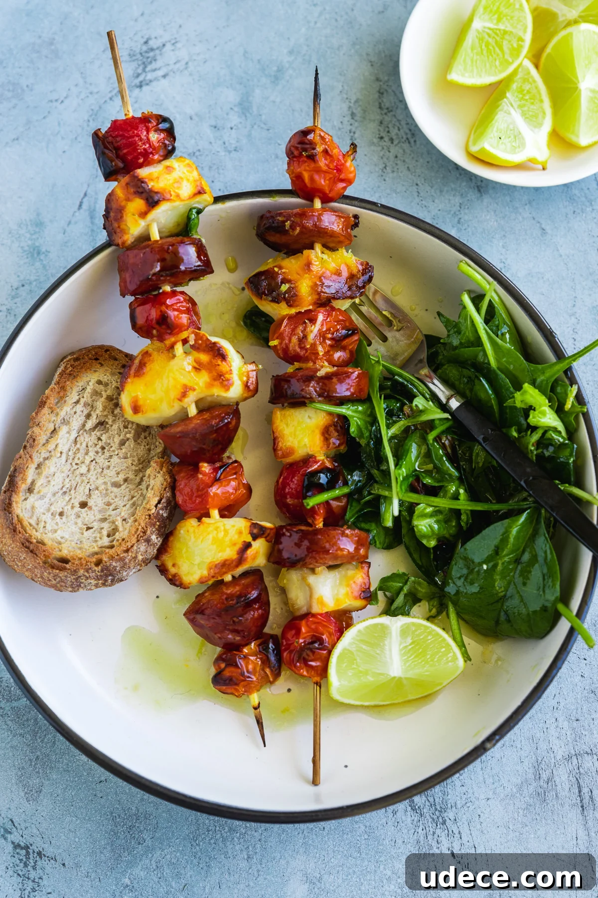 A dish of 2 halloumi kebabs with chorizo and tomatoes from above served with toasted bread, green salad leaves and lime wedges on the side