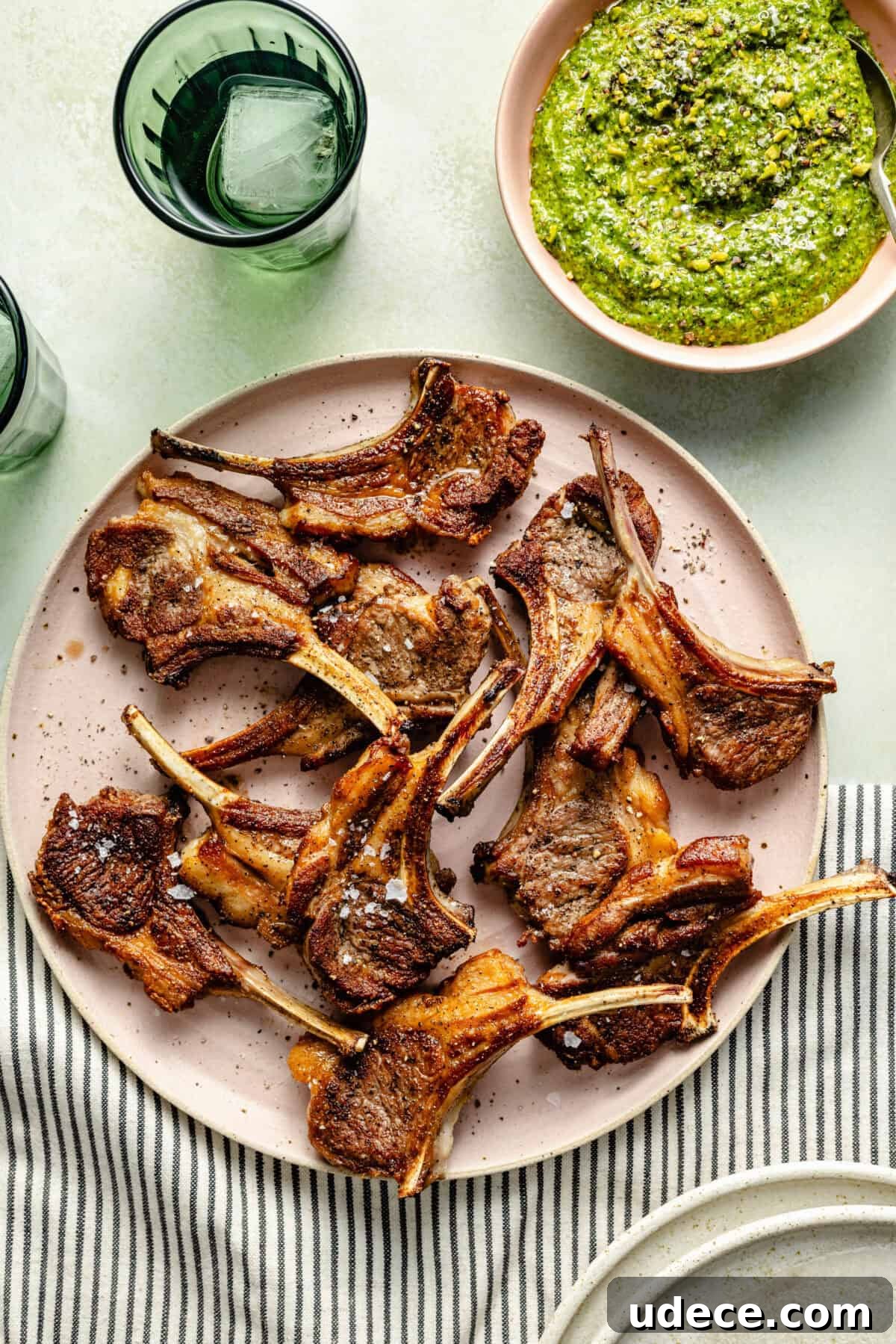 Grilled Lamb Chops with Fresh Arugula Pesto Recipe 4 a light pink plate filled with grilled lamb chops and a side bowl of arugula-pistachio pesto on a green background