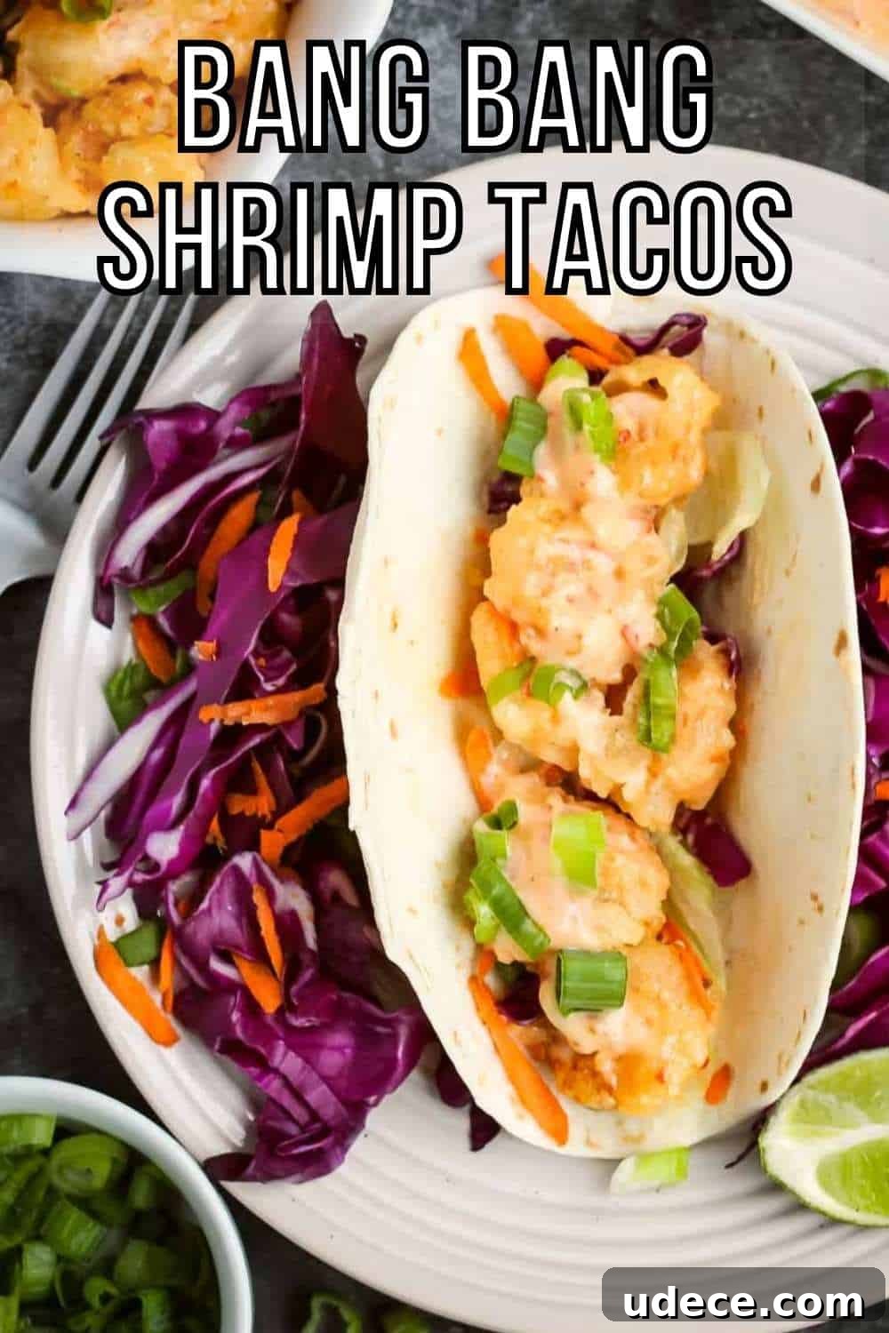 Overhead shot of one shrimp taco with shredded carrots and purple cabbage surrounding it and a lime wedge on the side of the white plate. On the black surface surrounding, there is more shrimp, sauce, and chopped green onions in white bowls as well as a fork and grey cloth.