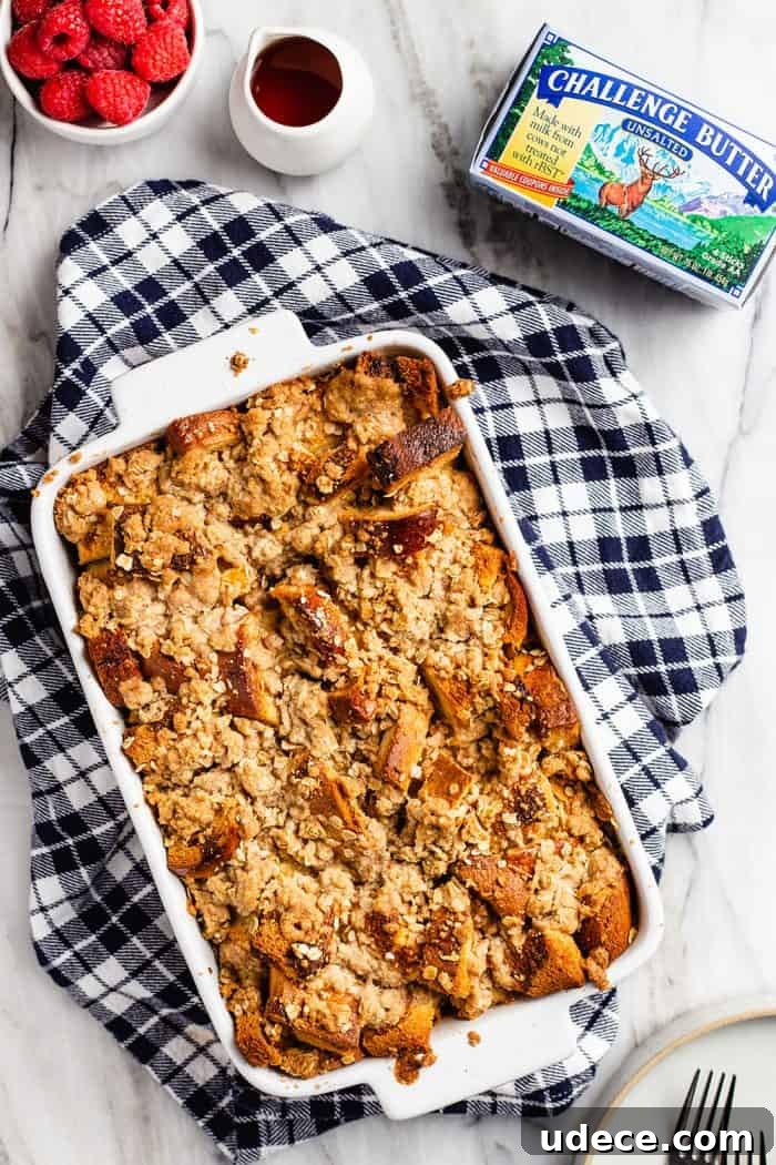 Overnight French Toast Casserole Recipe for Make-Ahead Brunch 9 Overhead view of overnight french toast casserole in a white baking dish set on a blue plaid dish towel. A bowl of raspberries, a pitcher of syrup, and a package of butter are nearby.