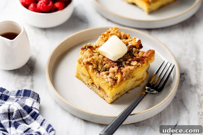 Overnight French Toast Casserole Recipe for Make-Ahead Brunch 10 Slice of french toast casserole topped with a pat of butter next to a fork on a white plate. A pitcher of syrup and a bowl of raspberries are in the background.