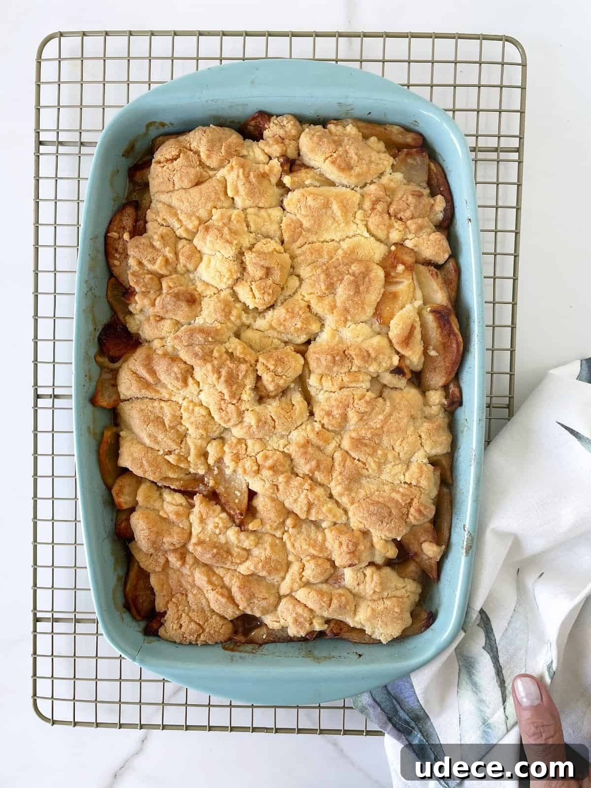 Caramel Apple Dump Cake Recipe — Quick Crowd-Pleasing Dessert 9 Baked apple dump cake in a blue dish on a wire rack. A kitchen towel. White marble surface.