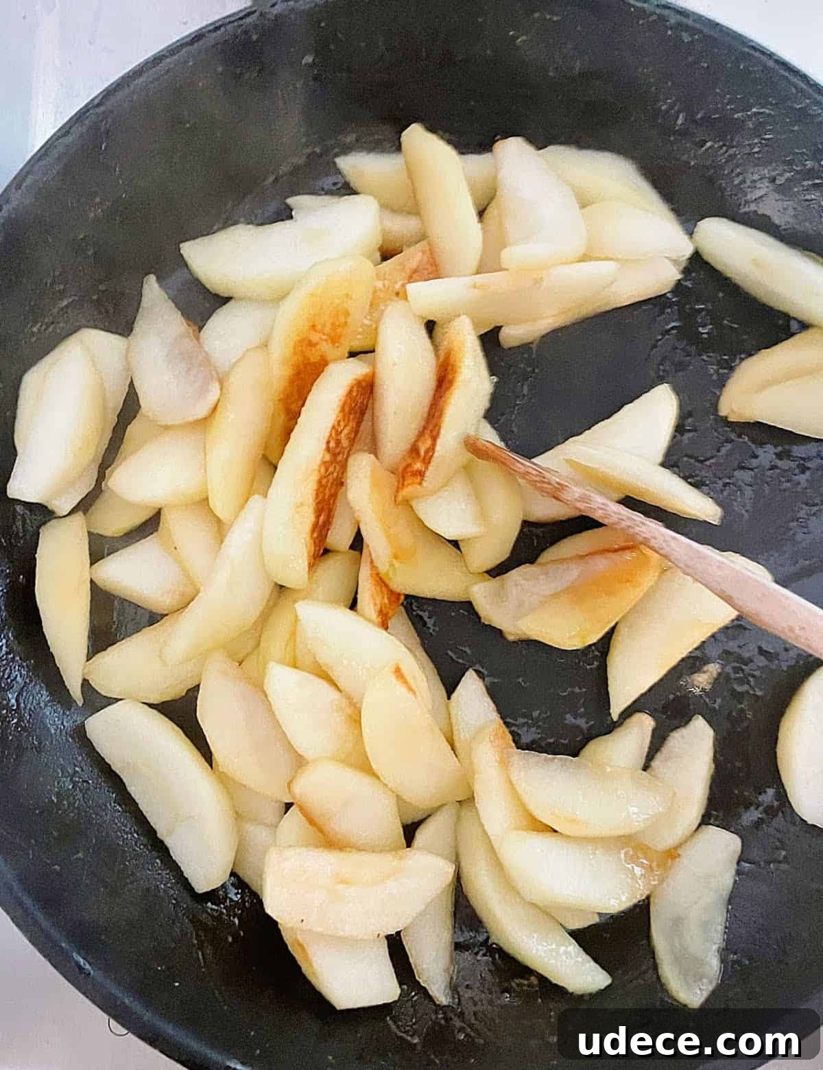 Caramel Apple Dump Cake Recipe — Quick Crowd-Pleasing Dessert 10 Cooking apple slices in a black skillet with a wooden spoon.