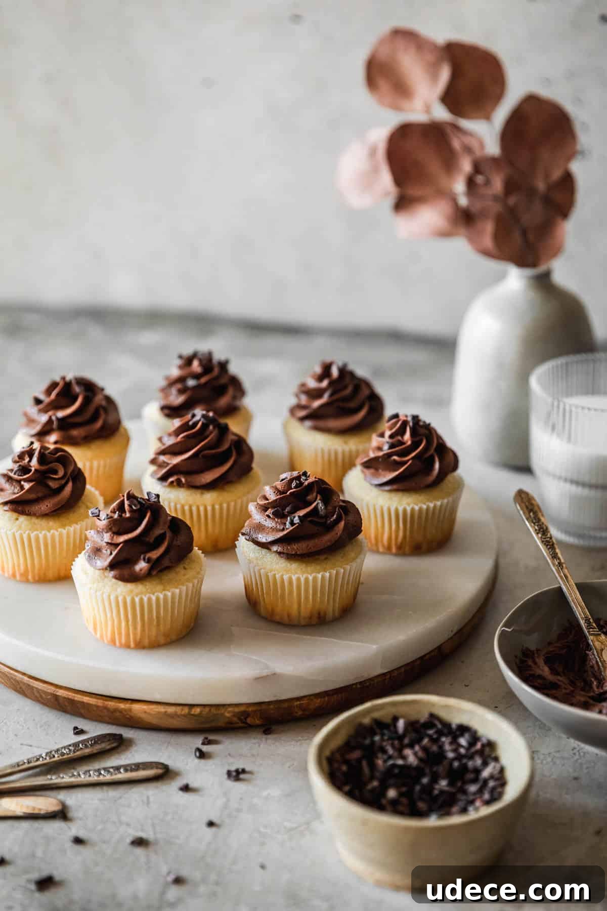 High-Altitude Yellow Cupcakes with Rich Chocolate Buttercream 13 A side image of a marble and wood tray of high altitude yellow cupcakes with chocolate buttercream on a grey table next to a grey bowl of frosting, white bowl of cacao nibs, white vase with brown blossoms, and cup of milk.