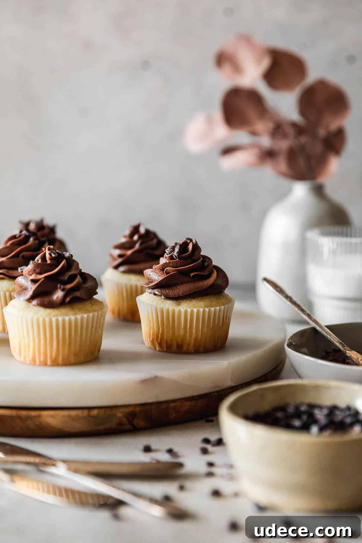 High-Altitude Yellow Cupcakes with Rich Chocolate Buttercream 14 A closeup side image of high altitude yellow cupcakes with chocolate buttercream on a marble and wood cake stand next to a grey vase with blossoms, a glass of milk, a grey bowl of frosting, and a white bowl of cacao nibs with a grey background.