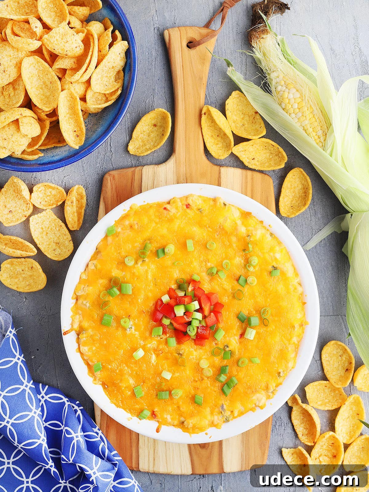 hot corn dip with cream cheese in a white pie plate garnished with green onions and red and green diced bell peppers. The plate rests on a wooden cutting board and a blue of corn chips sits to the side. Loose corn chips are sprinkled around.