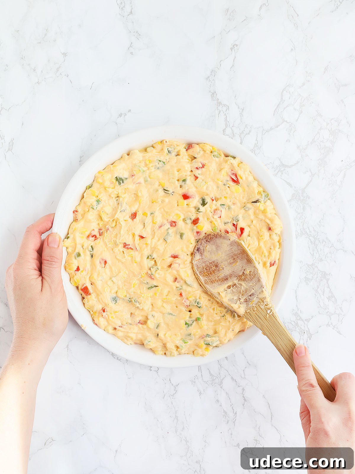 hand using a wooden spoon to spread the corn dip mixture into a white pie plate