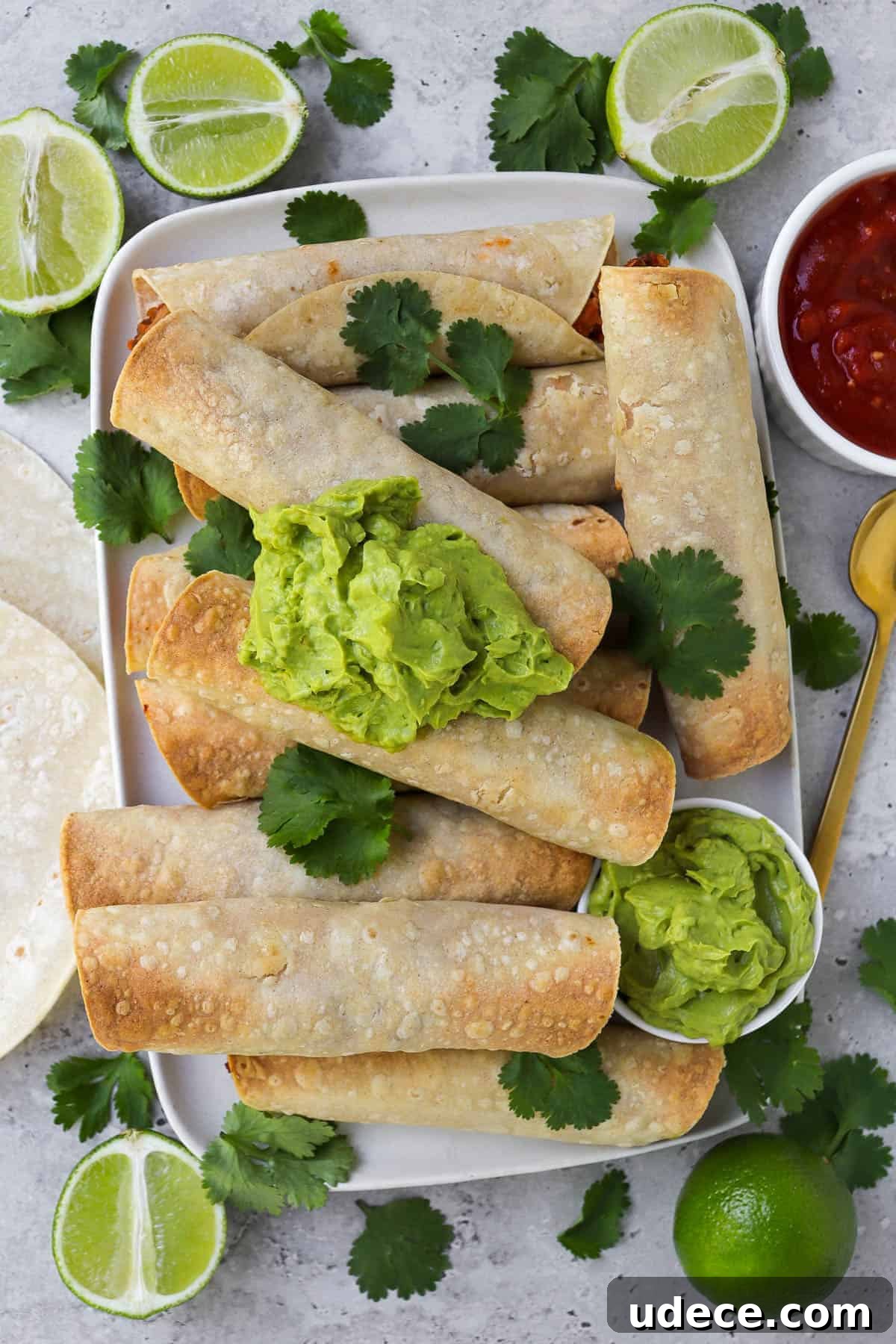 Vegan Lentil and Roasted Cauliflower Taquitos Recipe 2 Taquitos on platter topped with coriander leaves and guacamole. Fresh cut up limes, tortillas, salsa and gold spoon on the sides.
