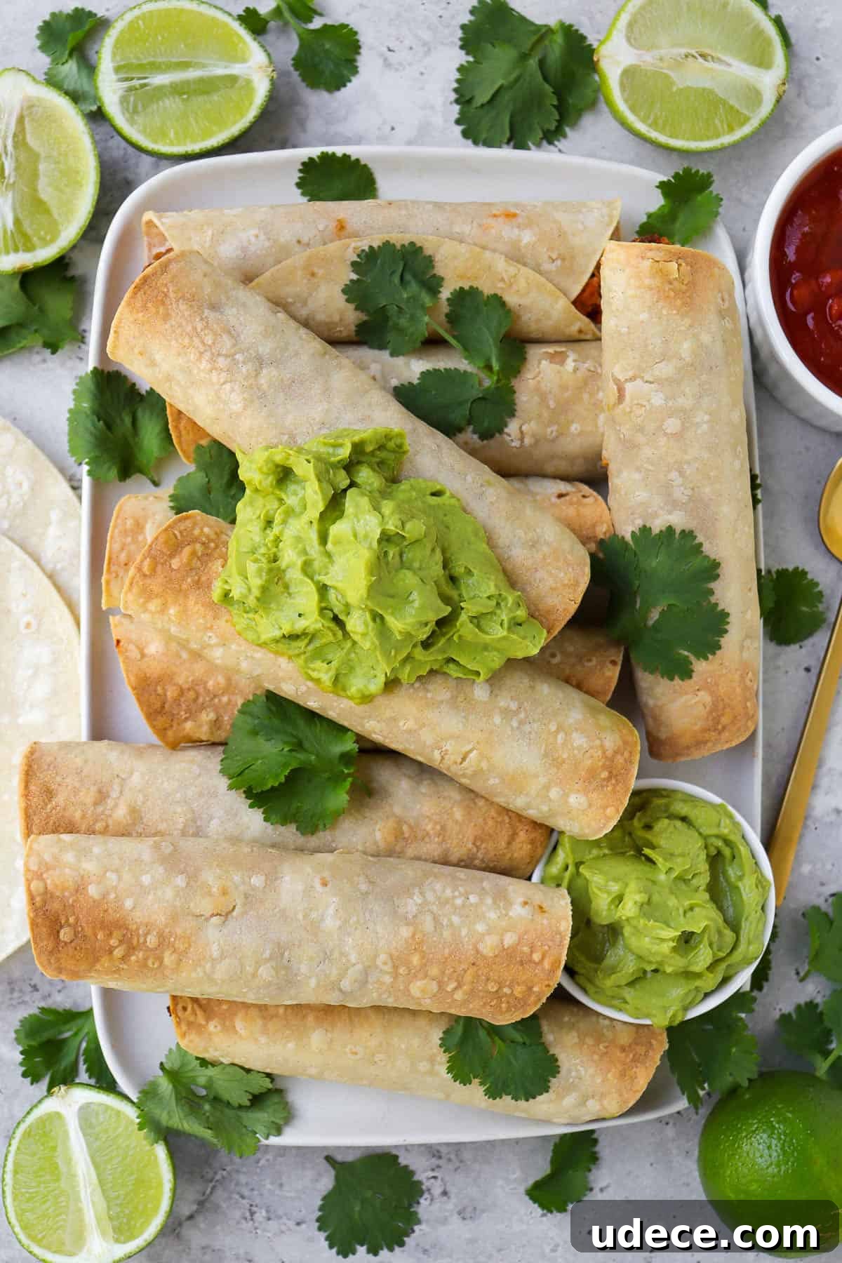 Vegan Lentil and Roasted Cauliflower Taquitos Recipe 7 Taquitos on platter topped with coriander leaves and guacamole. Fresh cut up limes, tortillas, salsa and gold spoon on the sides.