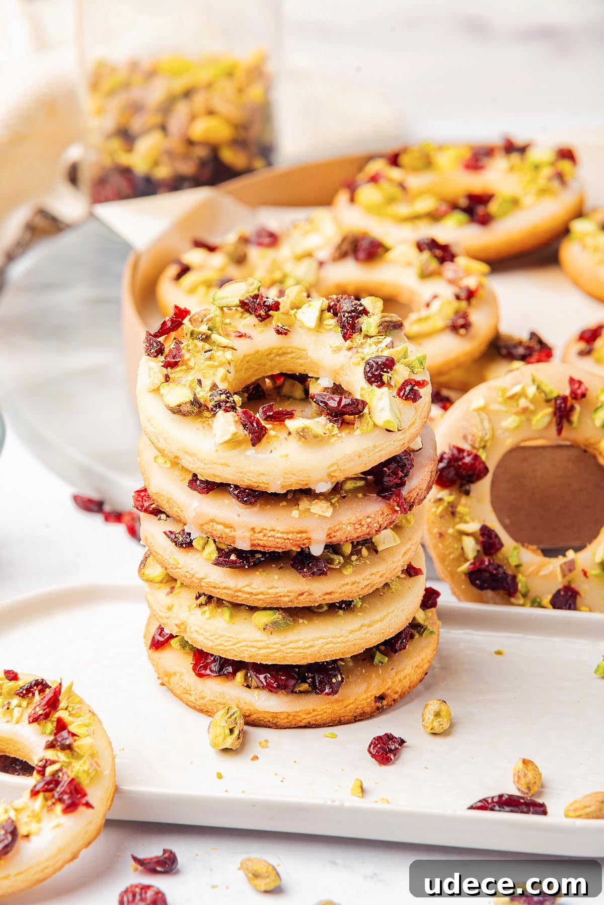 Cranberry Pistachio Holiday Wreath Cookies Recipe 11 A stack of cranberry pistachio wreath cookies topped with icing, pistachios, and dried cranberries rests on a white plate. Additional rings are in the background.