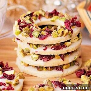 Cranberry Pistachio Holiday Wreath Cookies Recipe 12 A stack of cranberry pistachio wreath cookies topped with white icing, garnished with pistachios and dried cranberries, elegantly arranged on a wooden board.