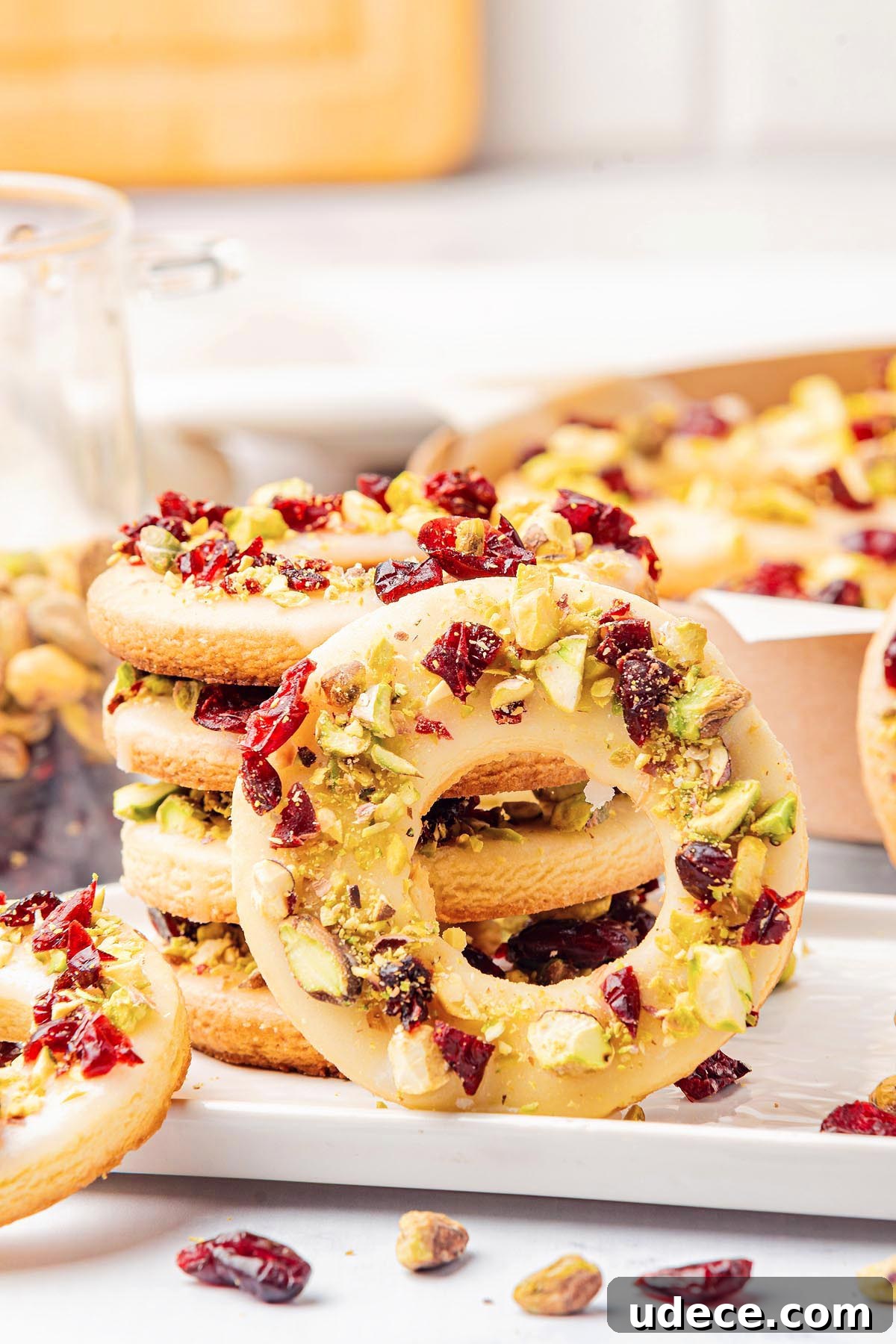 Cranberry Pistachio Holiday Wreath Cookies Recipe 4 A stack of white chocolate-covered donuts, reminiscent of cranberry pistachio wreath cookies, topped with chopped pistachios and cranberries on a white plate.