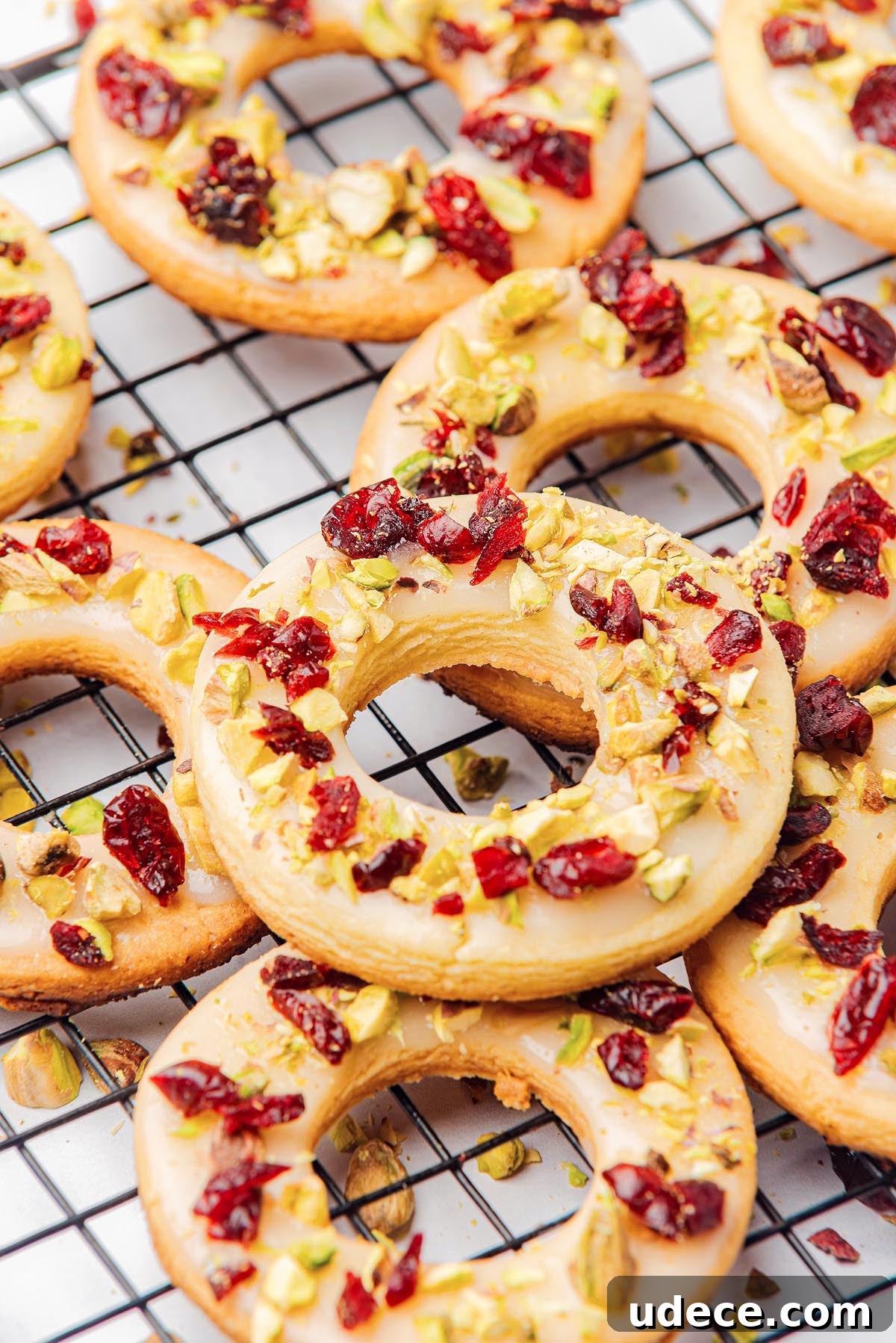 Cranberry Pistachio Holiday Wreath Cookies Recipe 8 Cranberry pistachio wreath cookies, delicately shaped like donuts, are adorned with white icing and topped with chopped pistachios and dried cranberries, elegantly arranged on a cooling rack.