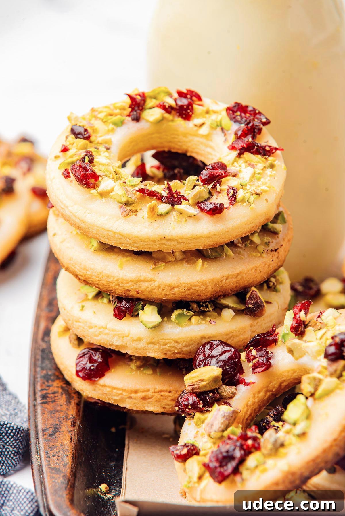 Cranberry Pistachio Holiday Wreath Cookies Recipe 9 A stack of four cranberry pistachio wreath donuts sits invitingly on a baking tray, topped with vibrant cranberries and crunchy pistachios, with a bottle of milk in the background.
