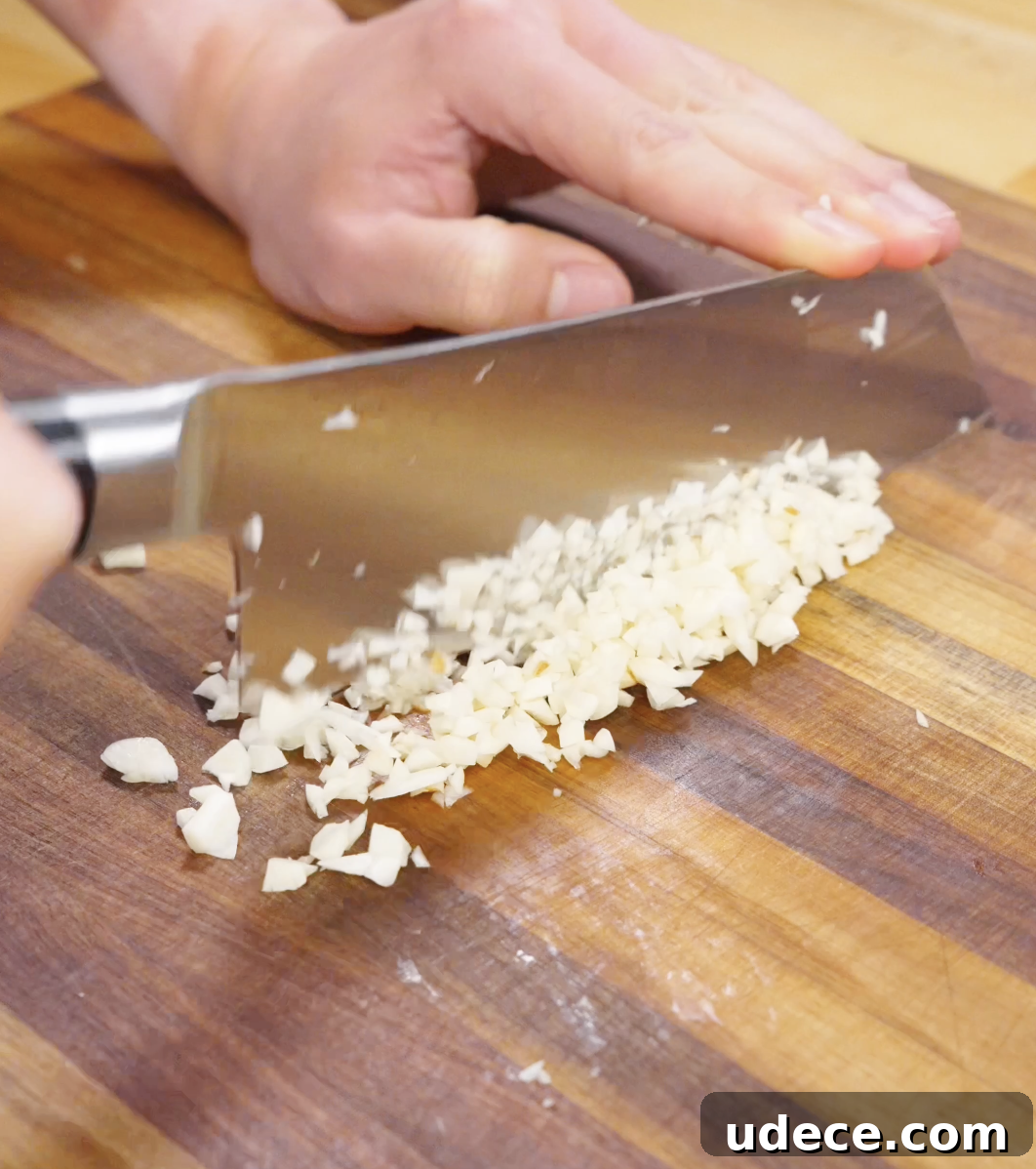 Garlic being finely minced on a wooden cutting board with a chef’s knife, preparing aromatic base for garlic beef fried rice.