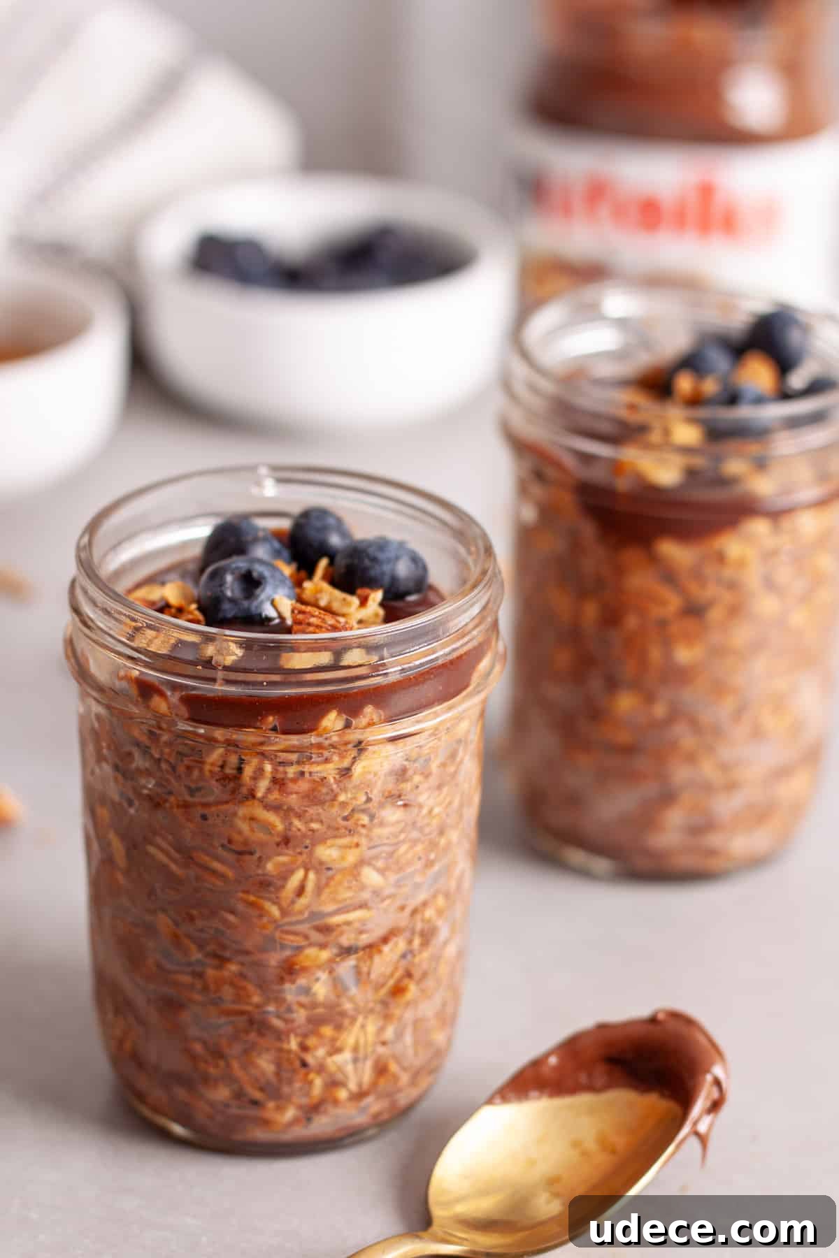 Nutella Overnight Oats Recipe for Creamy Chocolate Breakfast 2 A couple mason jars with Nutella overnight oats topped with blueberries and granola on a gray table.