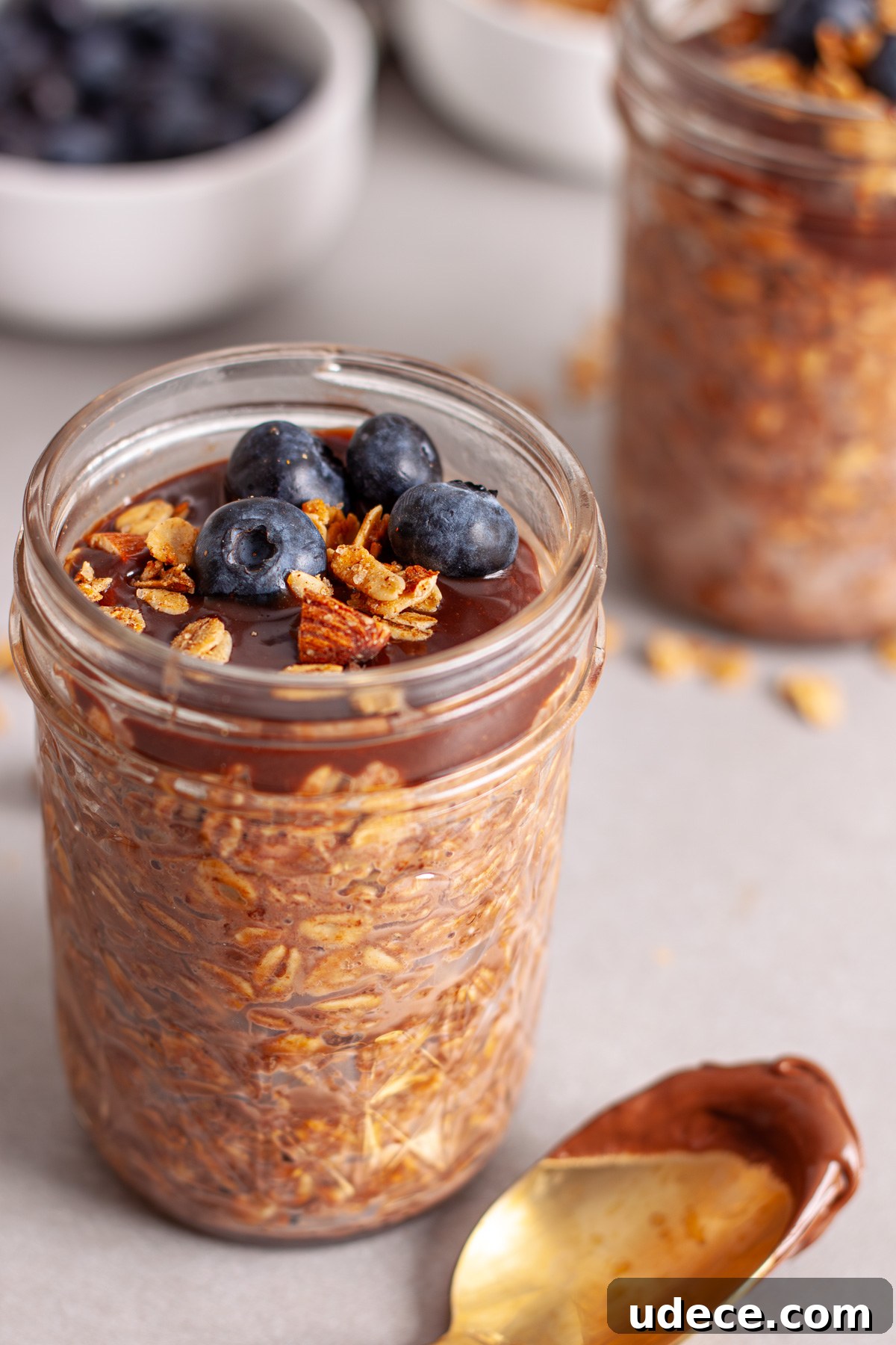 Nutella Overnight Oats Recipe for Creamy Chocolate Breakfast 10 A close up of a small mason jar of Nutella overnight oats topped with blueberries and granola on a gray table.