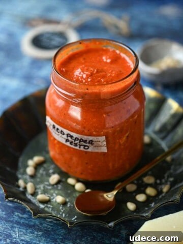 Red pepper pesto in a jar with grated cheese in the front on a blue surface.