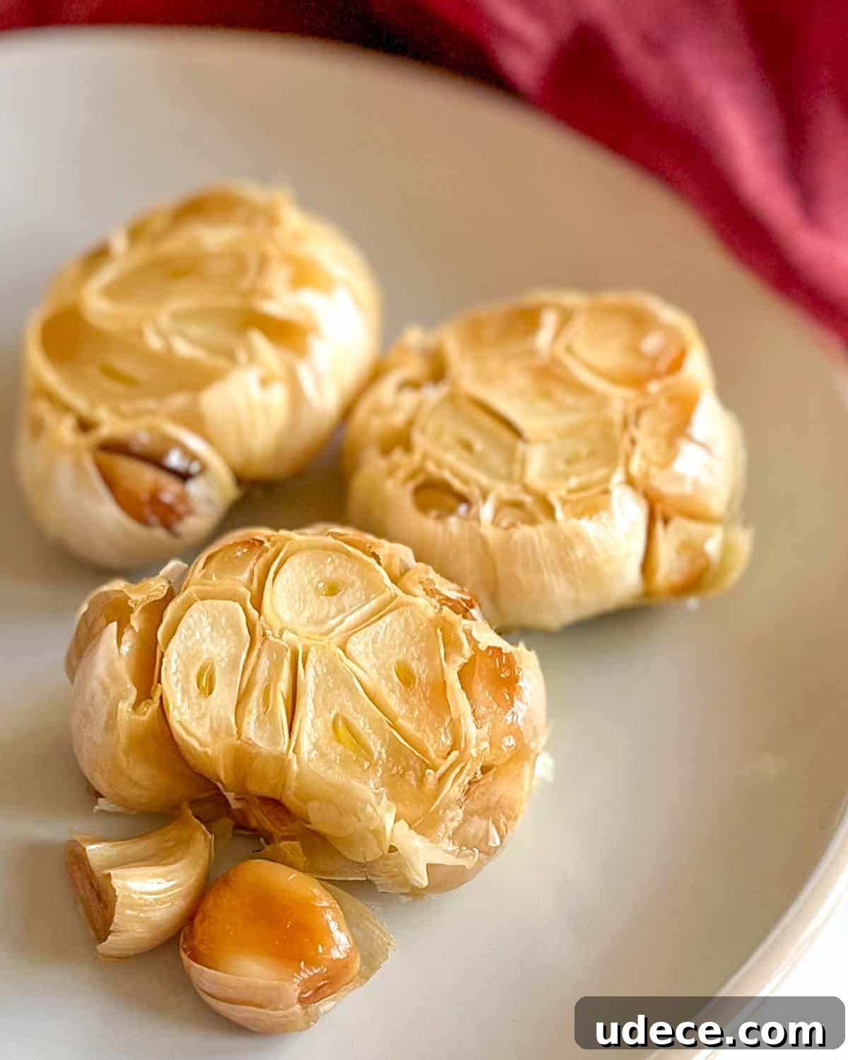 How to Roast Garlic Perfectly for Rich, Sweet Flavor 2 Three heads of roasted garlic sit on a white plate with a red linen in the background.