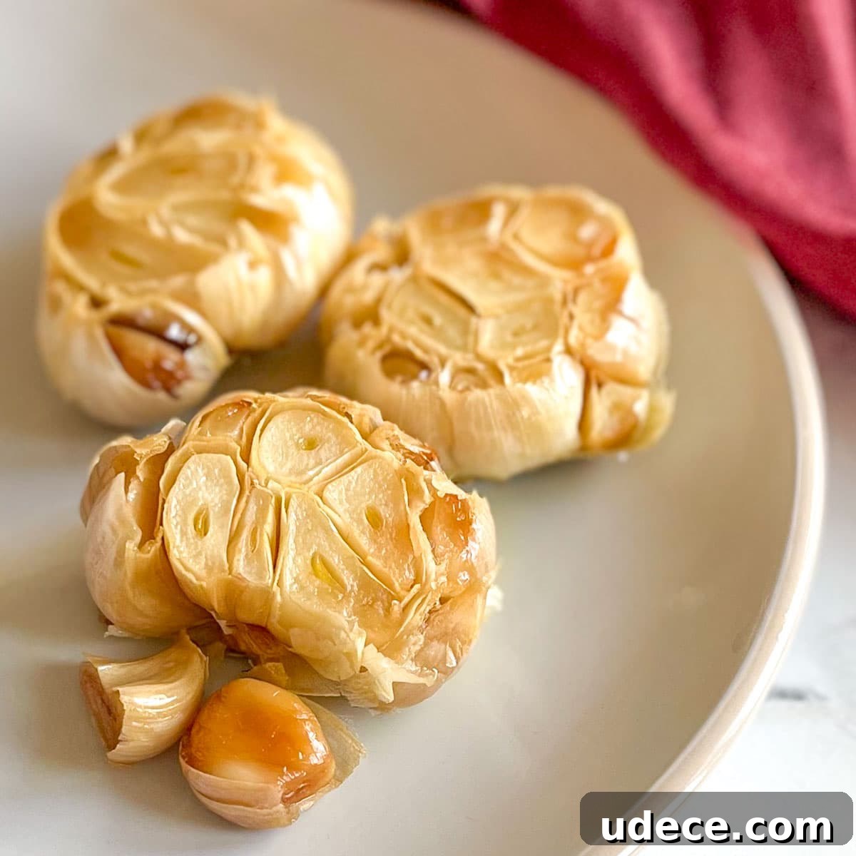 How to Roast Garlic Perfectly for Rich, Sweet Flavor 8 Three heads of roasted garlic sit on a white plate with a red linen in the background.