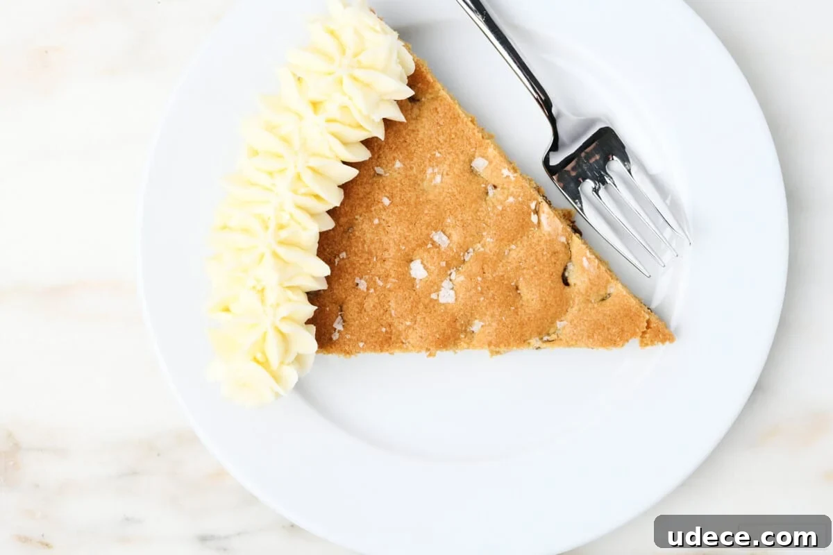 Overhead picture of slice of cookie cake with vanilla buttercream frosting and sprinkled with flake salt on a white dessert plate with fork.