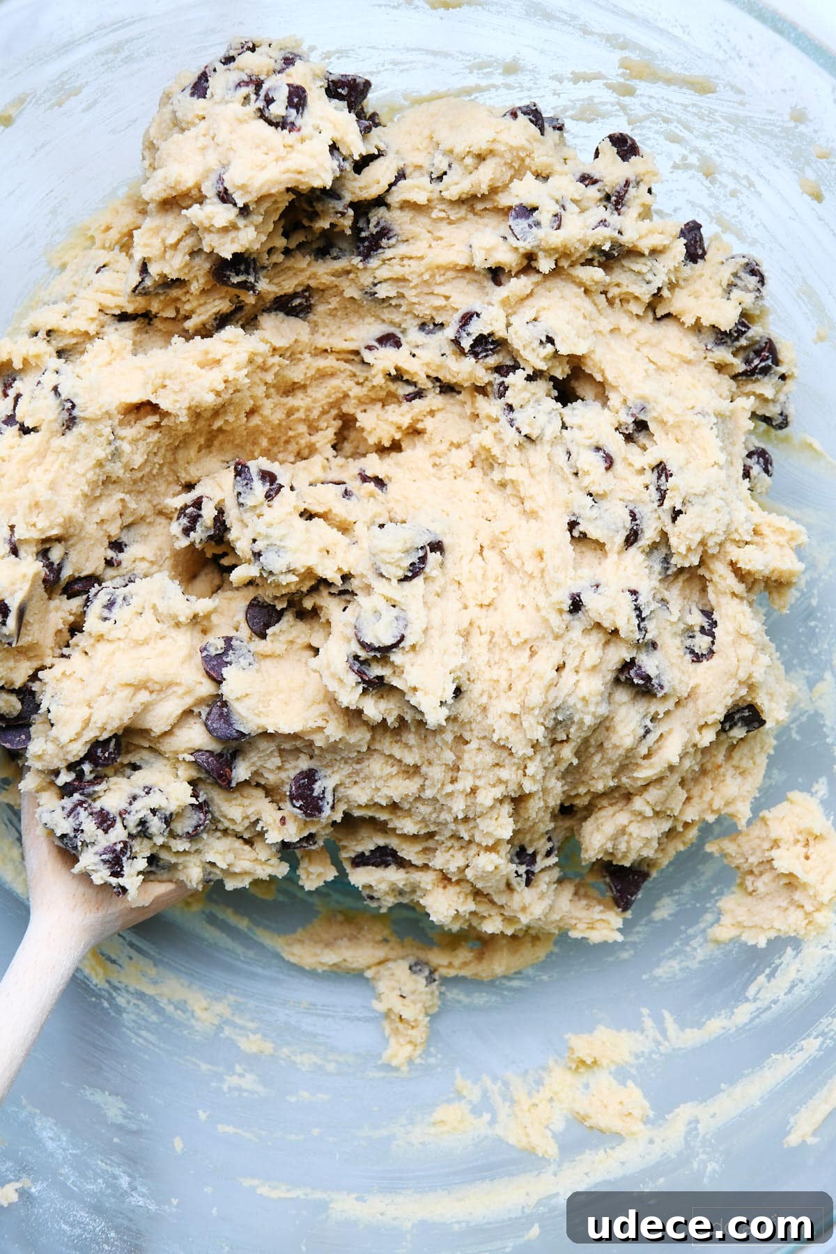 Chocolate chips blended into cookie cake dough in glass bowl with wooded spoon.