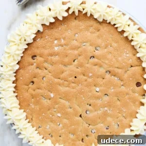 Vanilla buttercream frosting has been piped in star flower shapes around the edge of this chocolate chip cookie cake. Piping bag full of frosting is next to cake, all on marble countertop.