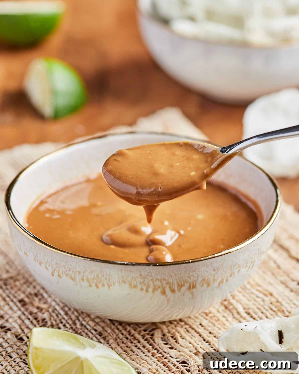 Photo of a spoonful of satay sauce lifted out of a bowlful with lime and crackers in the background.