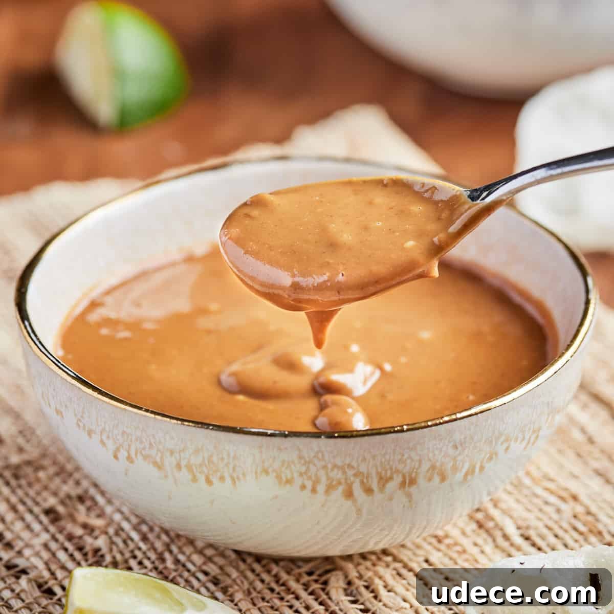 Square photo of a bowl of vegan satay sauce with a spoonful being taken out of it, set on a bamboo mat with lime wedges in the background.