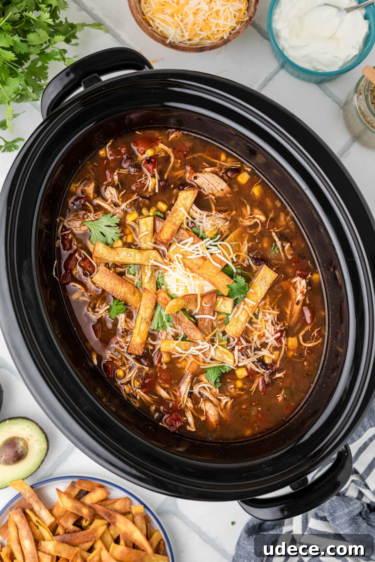 A black slow cooker filled with chicken tortilla soup, topped with some crispy tortilla strips, shredded cheese and sour cream.