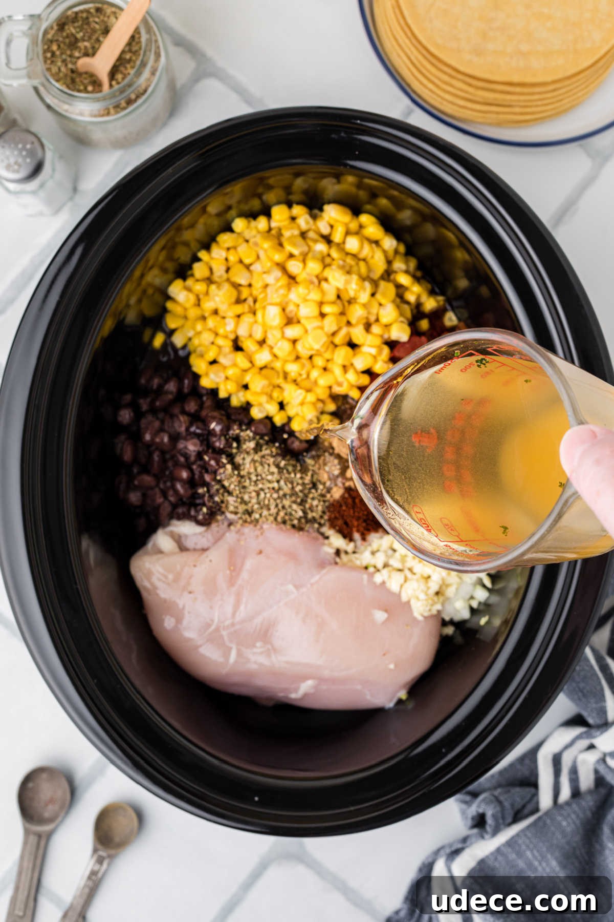 A black crock pot with ingredients, and chicken broth being poured in.