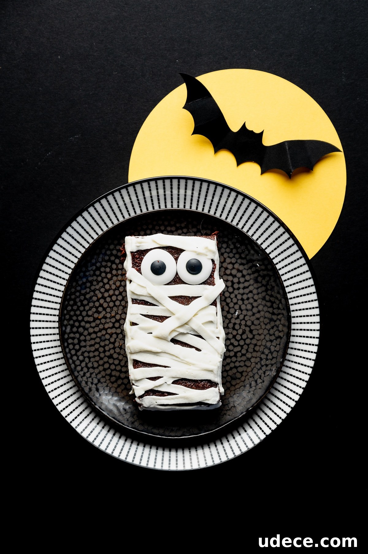 Sugar free brownie with googly eyes and frosting applied in strips to look like bandages on a black background with a bright yellow paper moon and paper bat